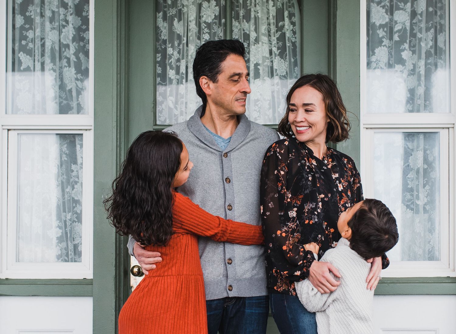 Happy family of four smiling together on a front porch, parents embracing two young children.
