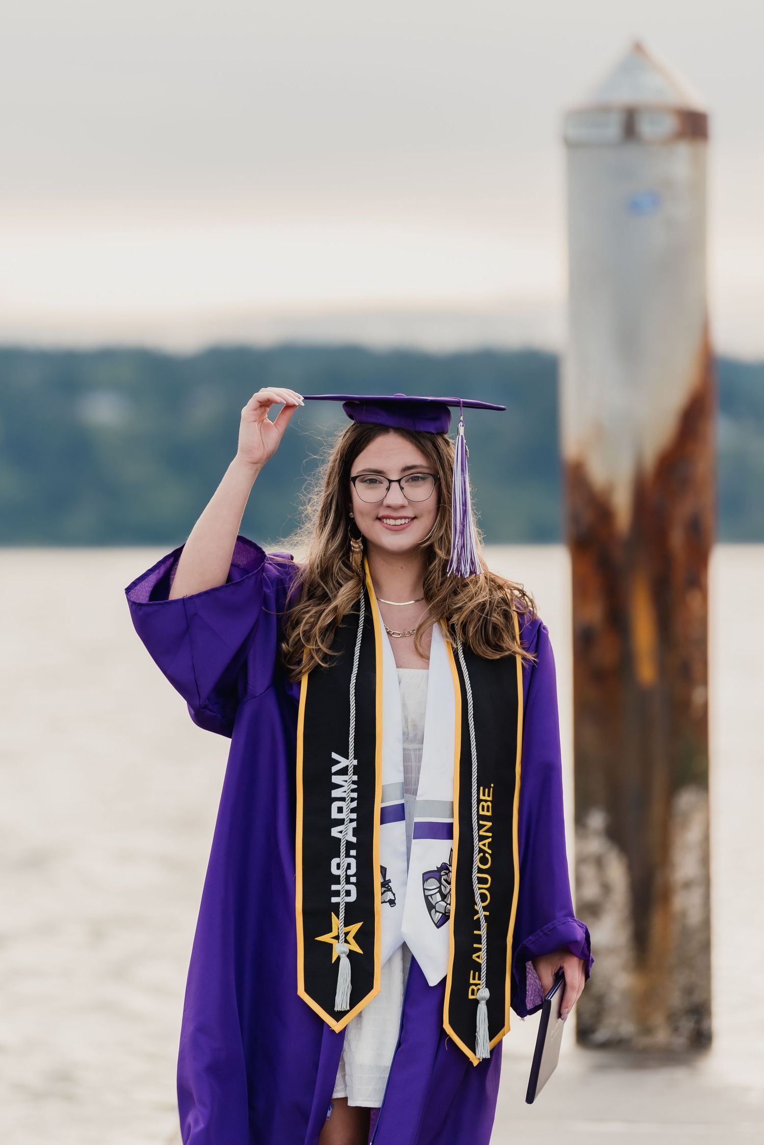Graduate in purple cap and gown with gold honor cords poses by waterfront wooden pilings at sunset.