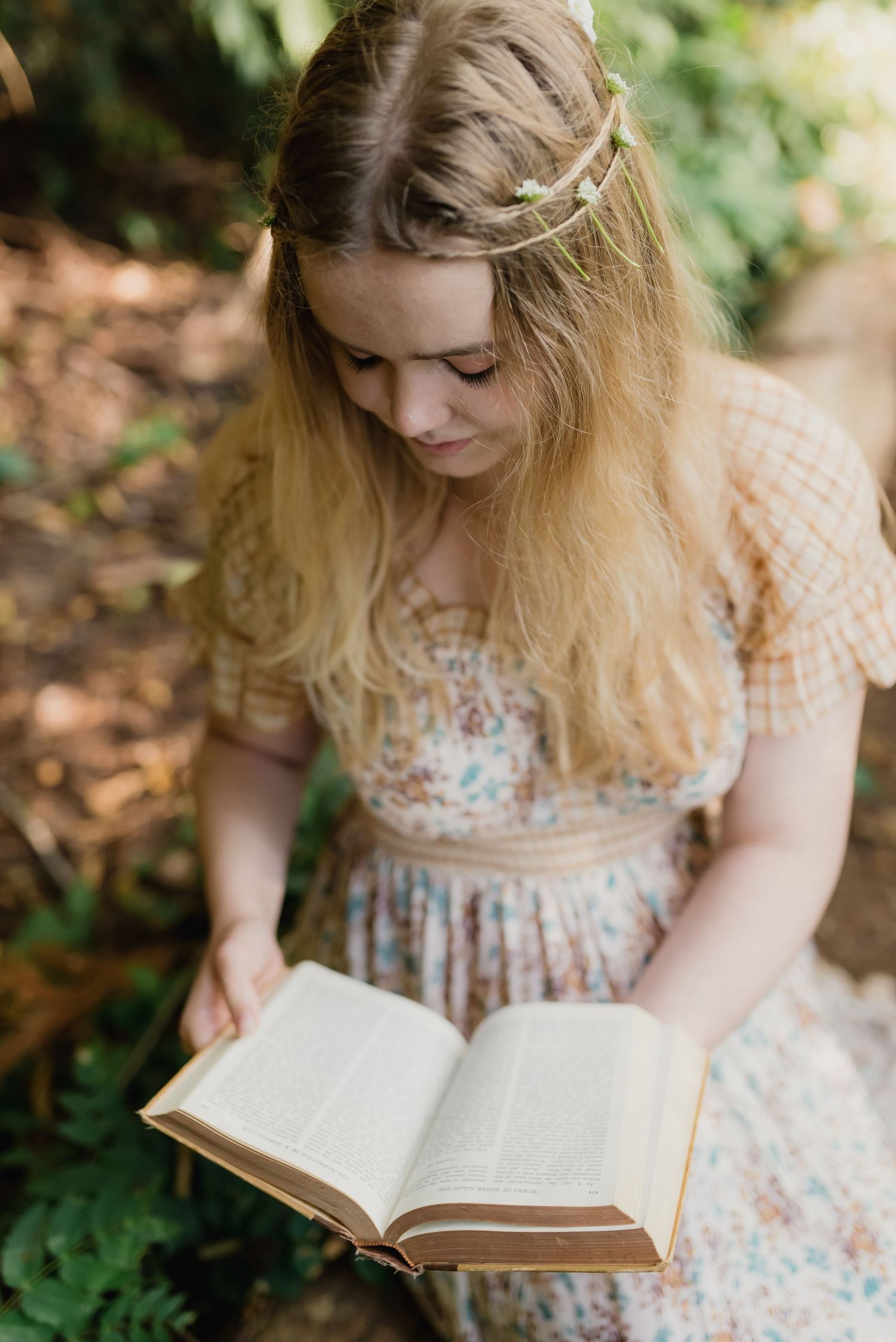 A reader in a floral dress with braided hair sits outdoors reading a book among greenery and fallen leaves.