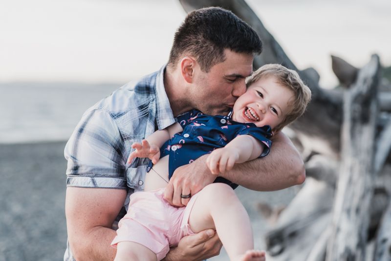 A father at the beach holds his toddler son and kisses his cheek while the boy giggles.