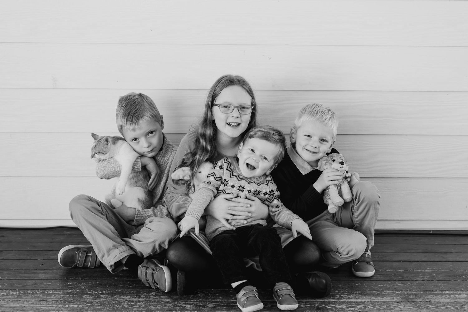 A black and white portrait of a group sitting together on a wooden floor against a light colored wall.