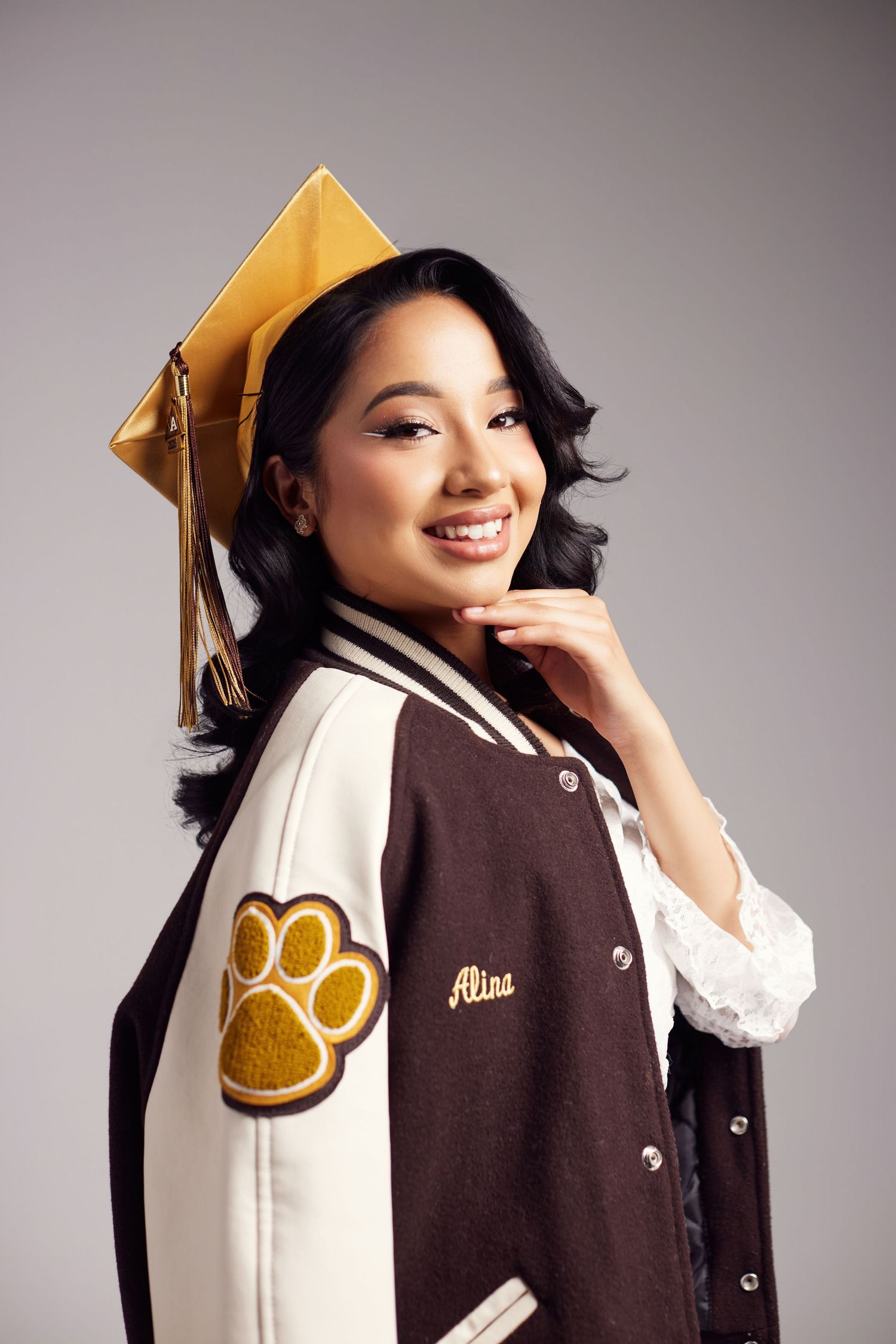 Smiling graduate wearing gold cap and brown varsity jacket with paw print patch and name Alina embroidered.