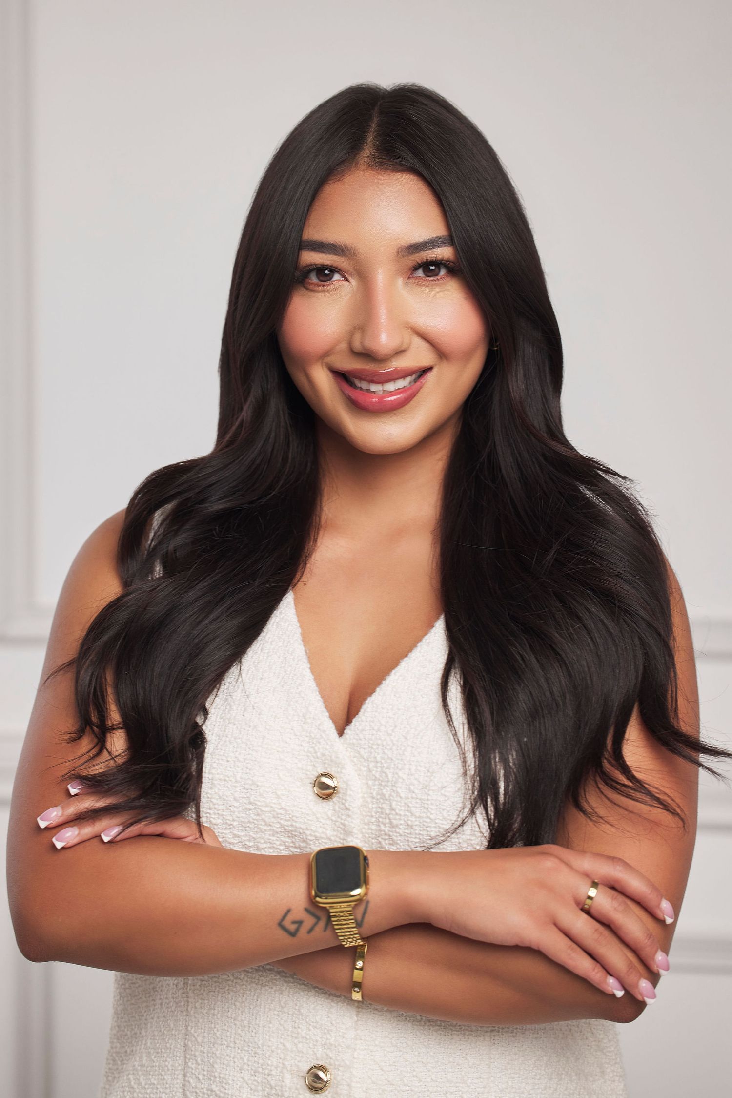 Professional portrait of someone with long dark hair wearing a white sleeveless dress and gold watch, smiling warmly.