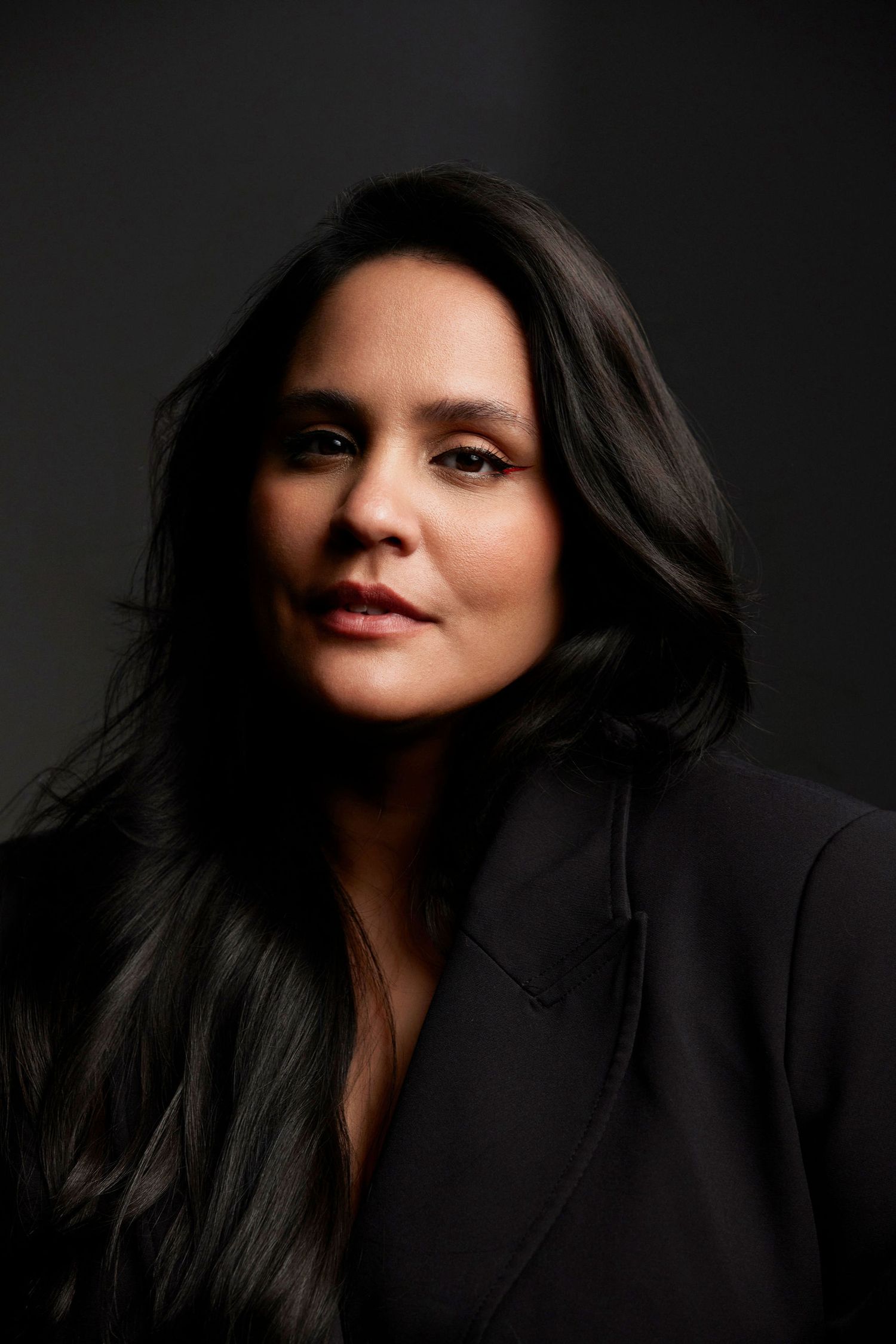 Woman with long dark hair wearing a black blazer poses against a dark background in a professional portrait.