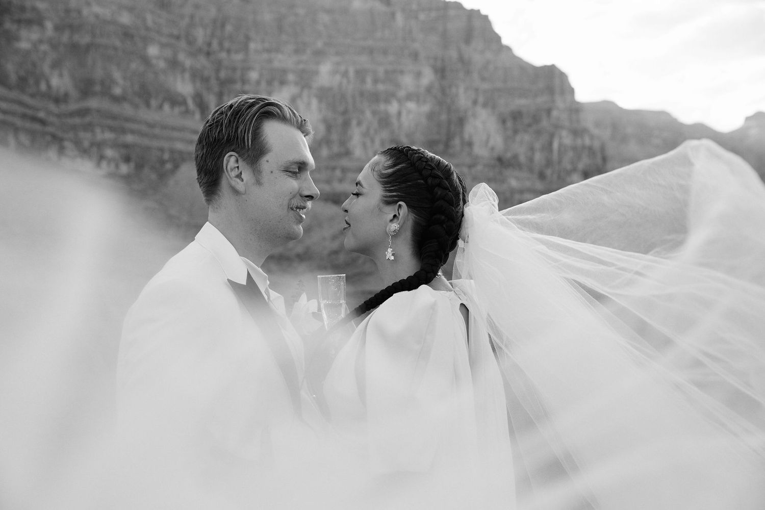 A romantic black and white portrait of newlyweds with a flowing veil against dramatic canyon cliffs.