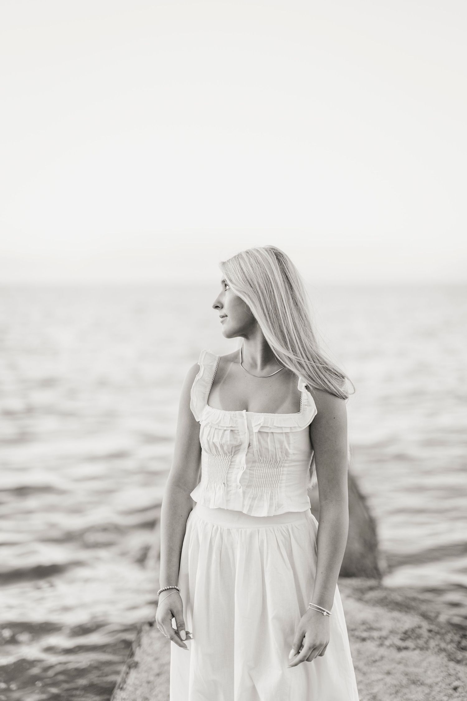 A person in a white sundress stands near the ocean shore in a serene black and white photograph.