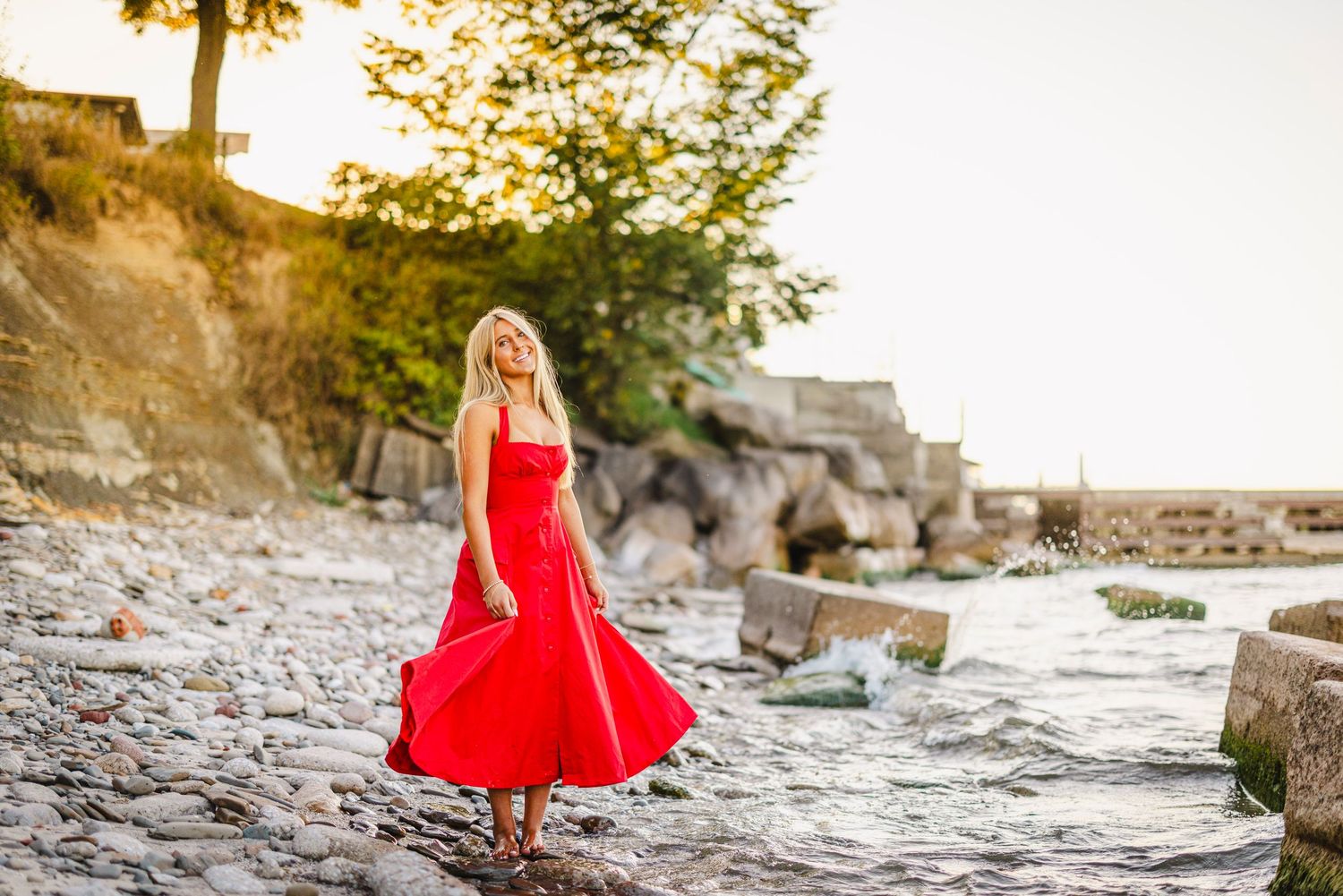 A flowing red dress catches the breeze along a rocky shoreline during golden hour sunset.