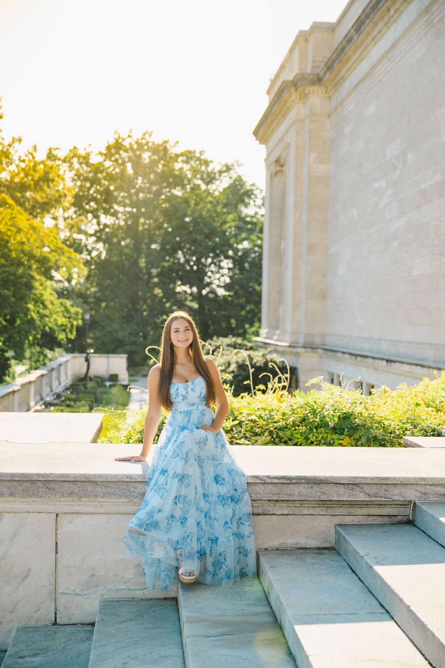 A person in a flowing blue floral dress poses gracefully on stone steps at sunset.