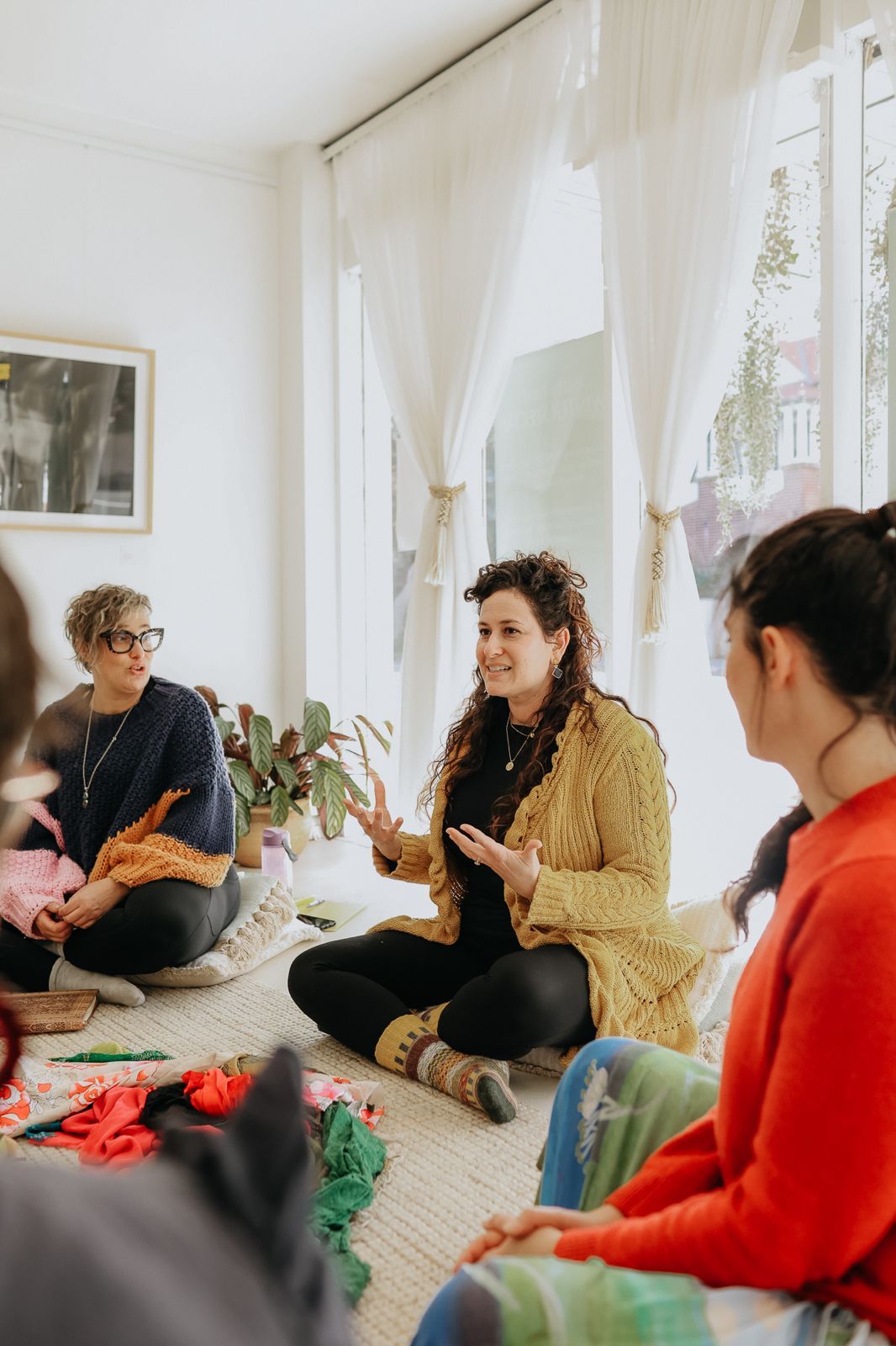 Women sitting in a circle on the floor during a creative workshop in a bright, plant-filled room.