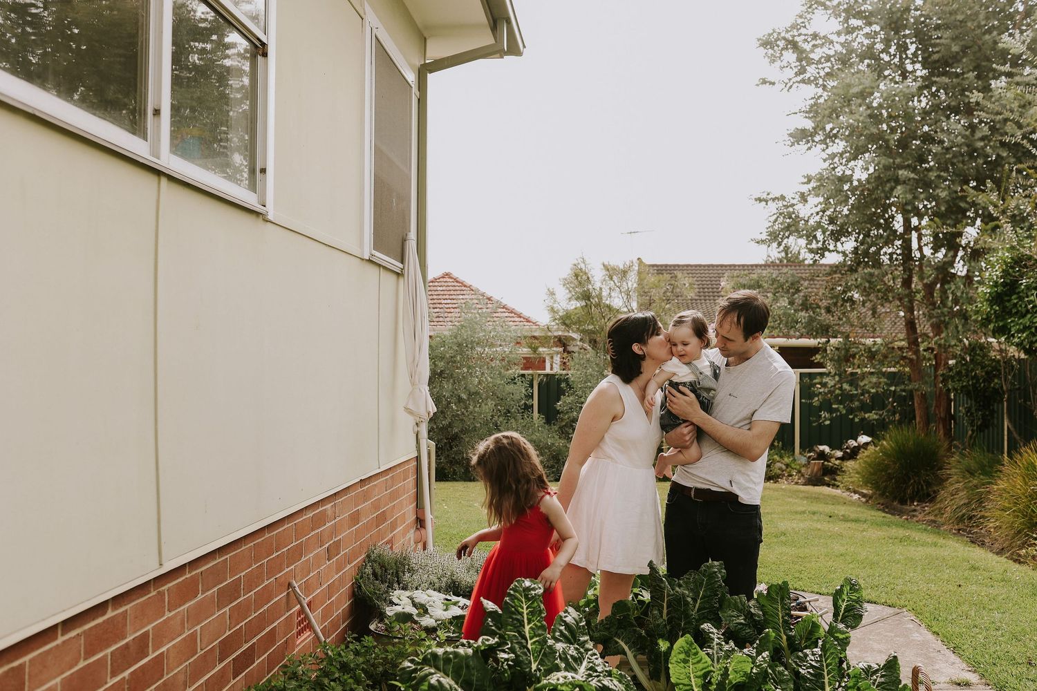Family of four enjoying time together in a lush backyard garden beside a brick home on a cloudy day.