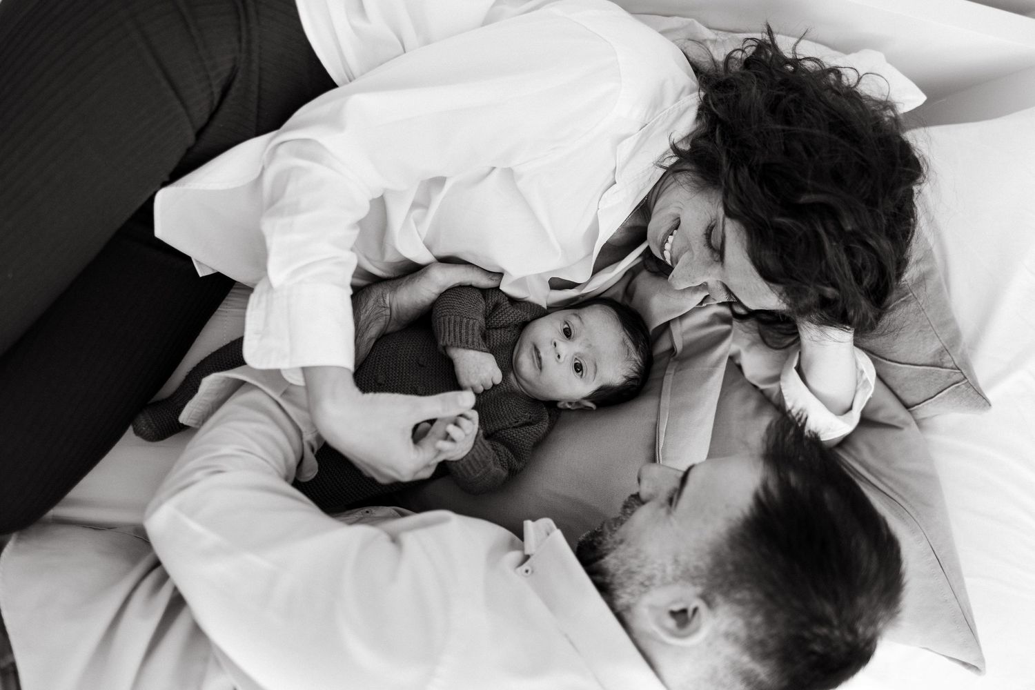 Black and white photo of parents lying on bed with newborn baby between them, sharing a tender family moment.