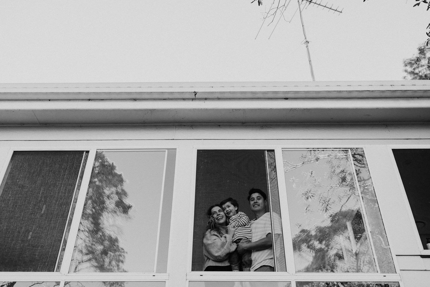 Black and white photo of a couple embracing and smiling inside a window of a mid-century modern home.