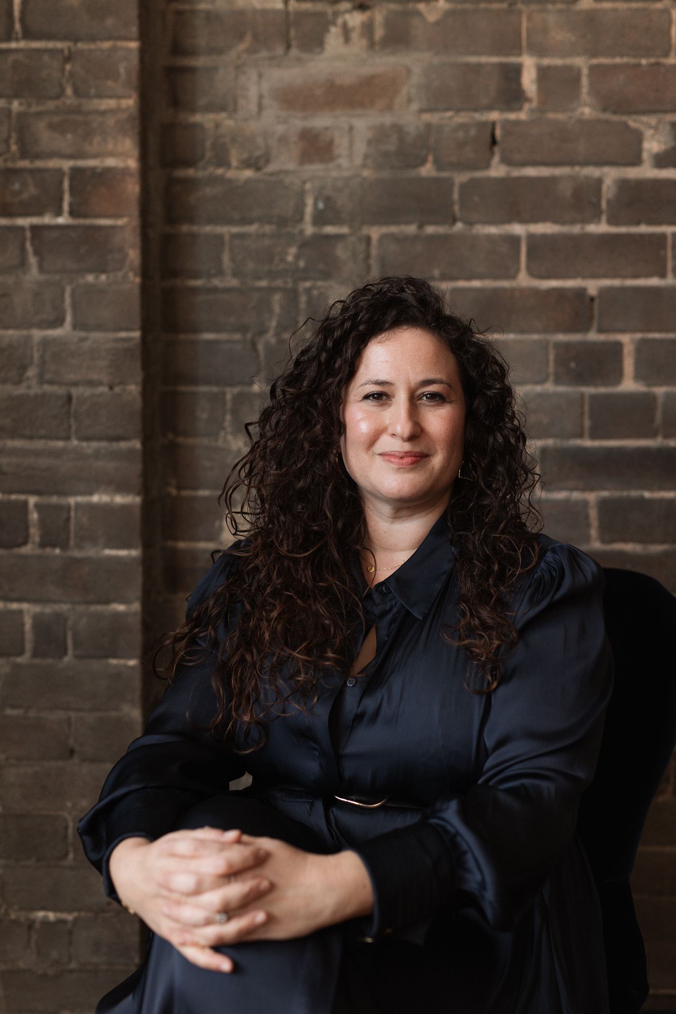 Woman with curly dark hair smiling, wearing a black satin blouse, seated against a rustic brick wall.