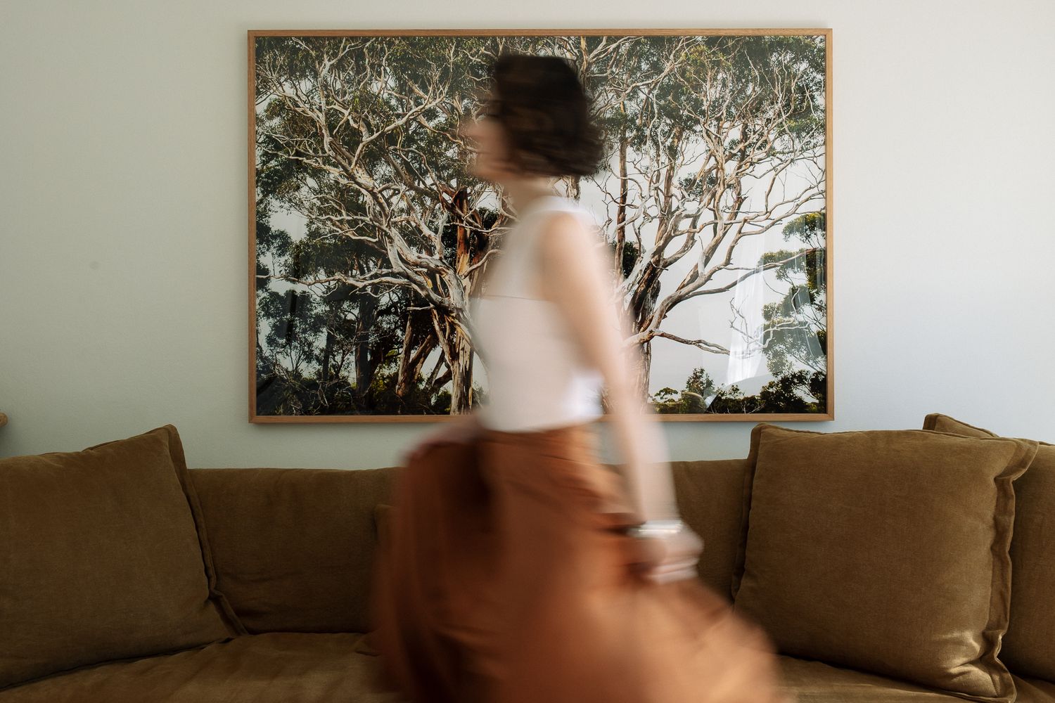 Woman in white top and rust skirt walks past a large tree painting above an olive green sofa.