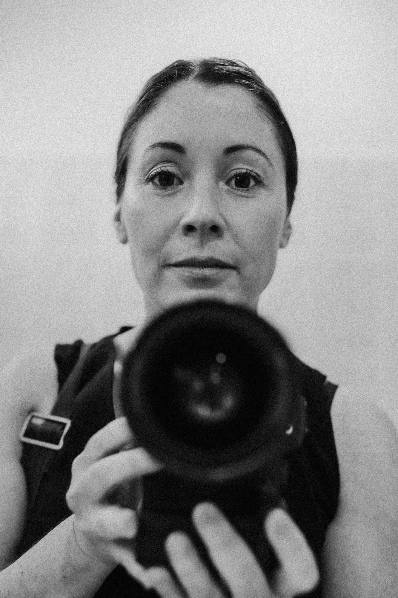 Black and white self-portrait of a woman holding a DSLR camera up to the lens in a mirror reflection.