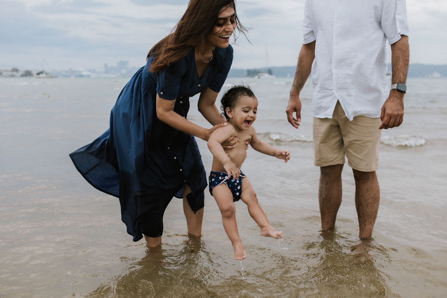 Family enjoying a beach day, parents playing with toddler in shallow ocean waves on a sunny day.