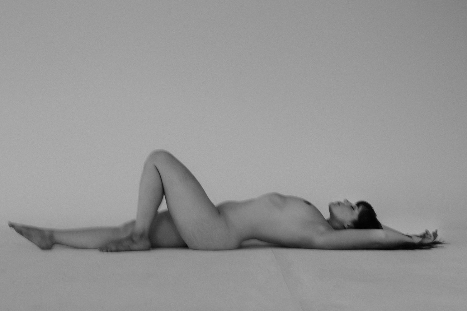 Black and white fine art nude photograph of a woman reclining gracefully on a light background.