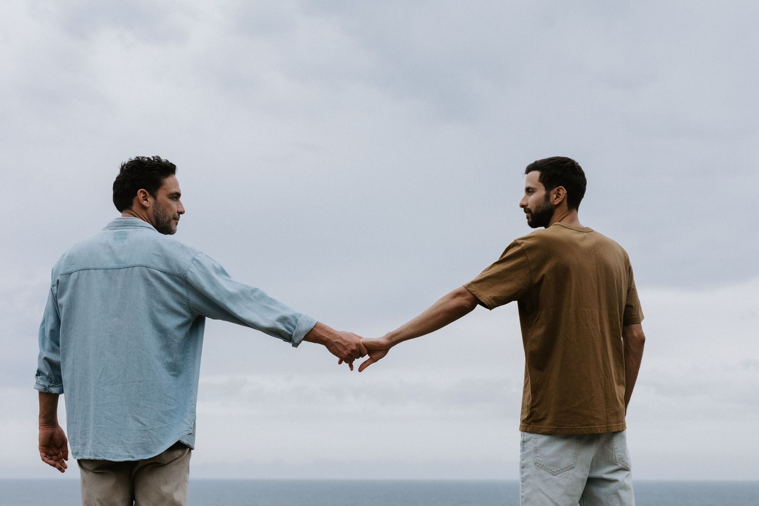 Two men reaching toward each other holding hands outdoors with an overcast sky and ocean in the background.