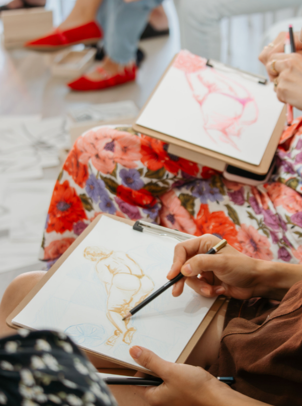 Artists sketching figure drawings in sketchbooks during a life drawing class, wearing floral dress.