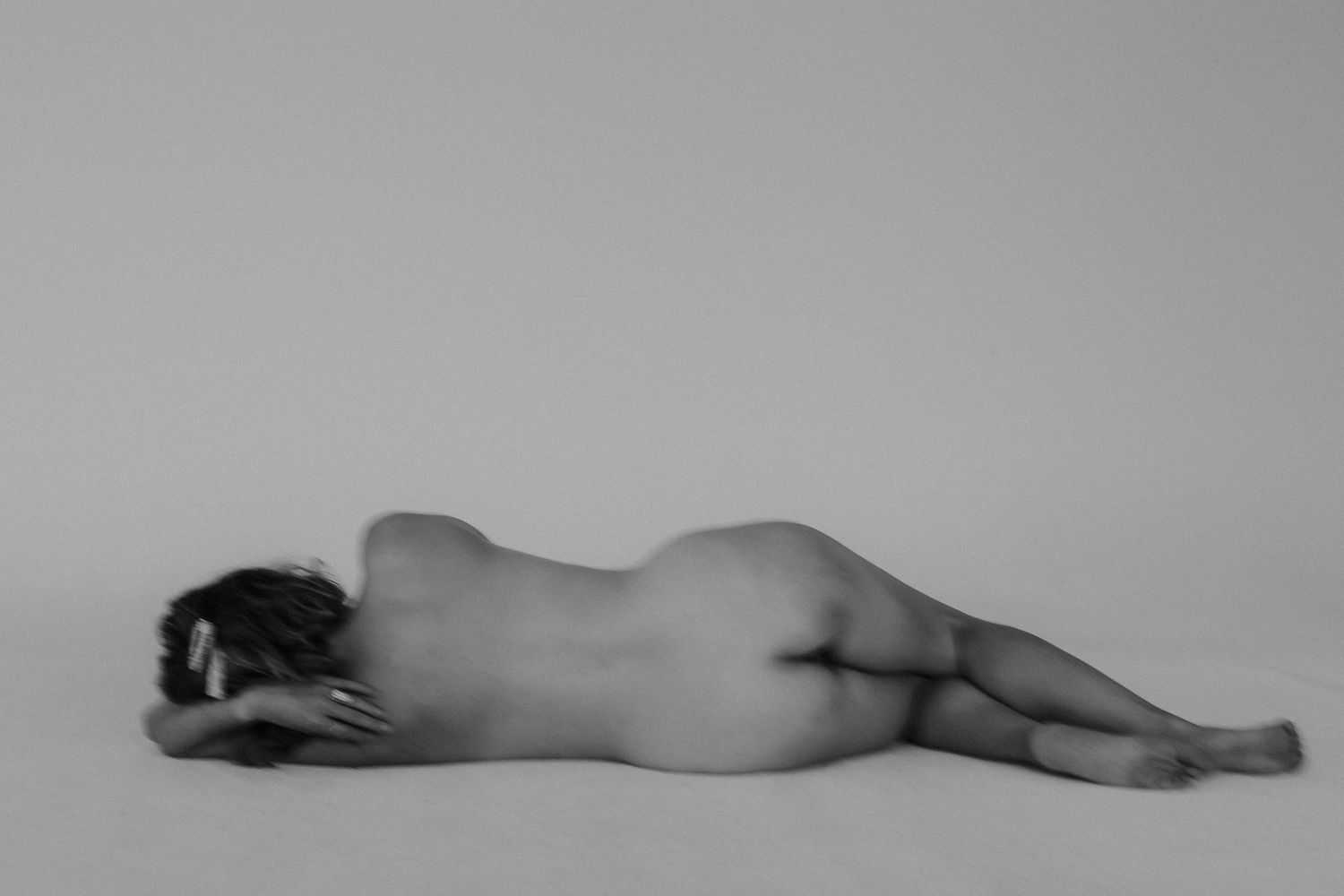 Black and white fine art nude photograph of a person lying on their side in a minimalist studio setting.