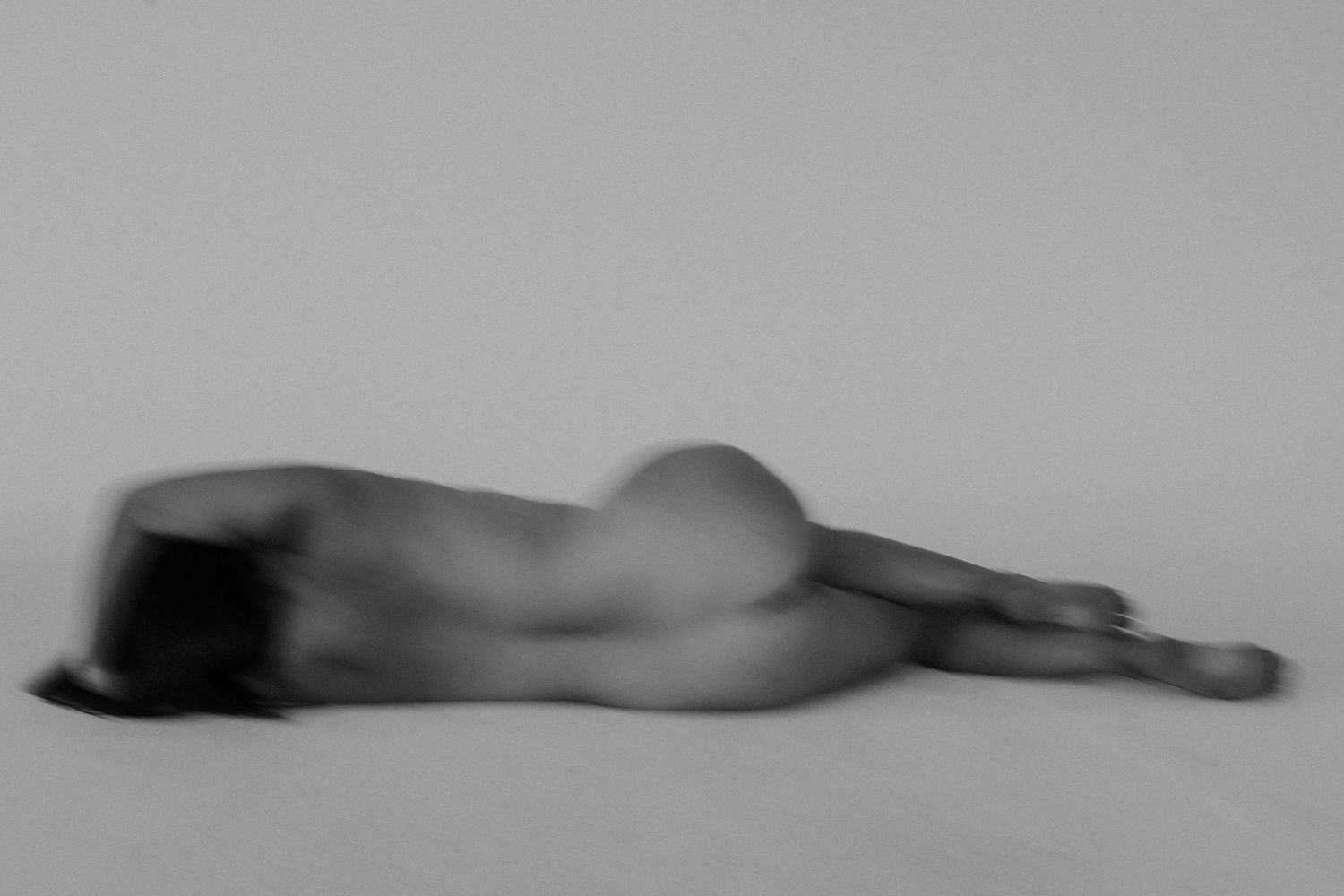 Black and white fine art photograph of a nude figure lying gracefully on a light surface with soft motion blur.