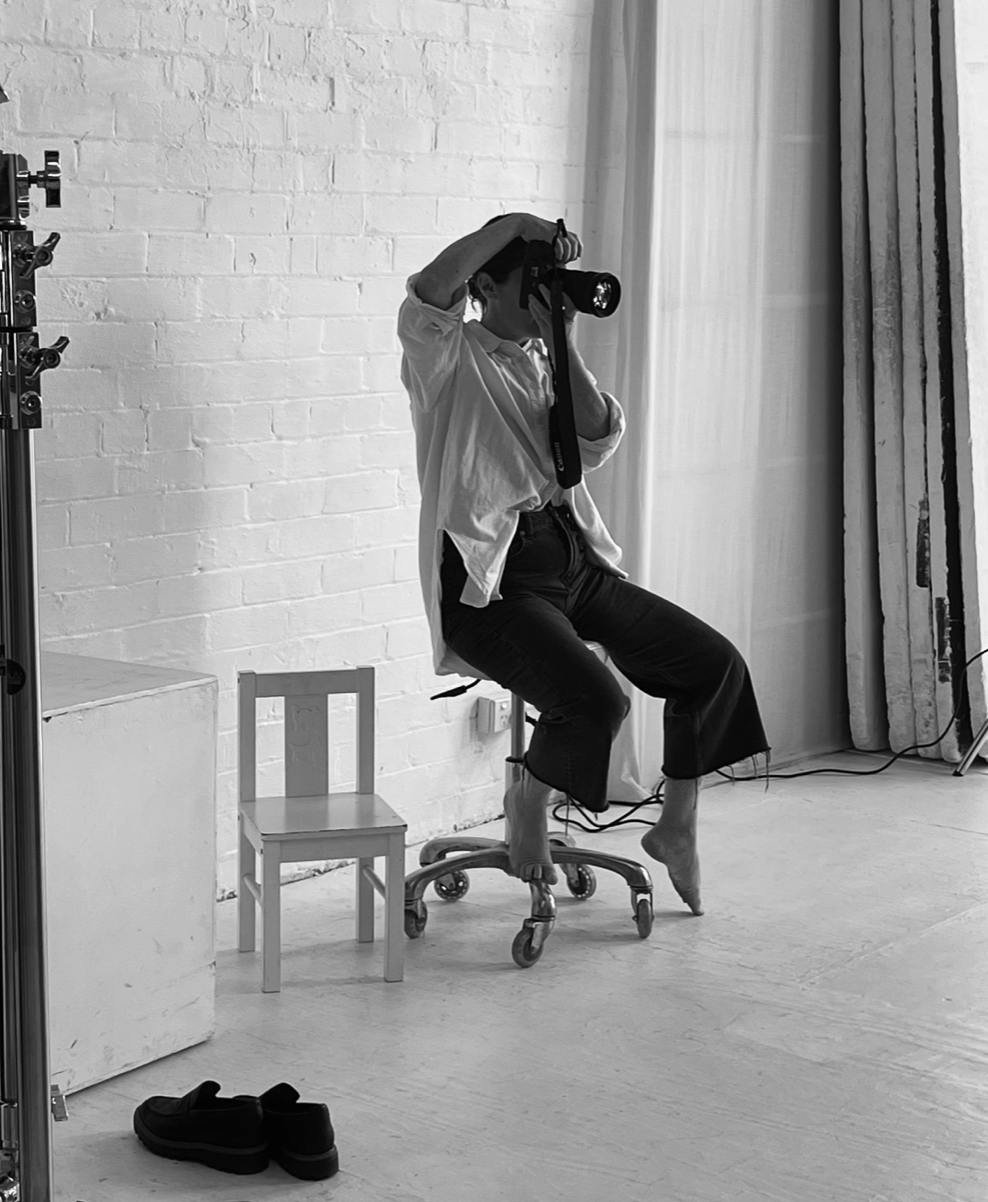 Black and white photo of a person sitting on a office chair holding a camera in a photography studio.