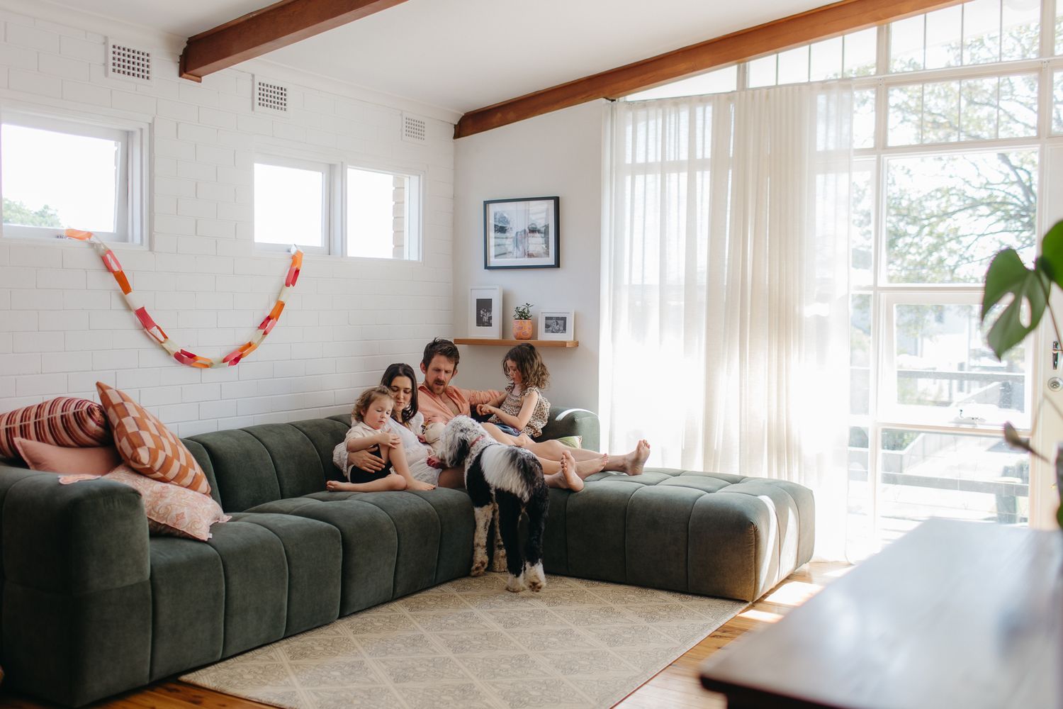 Family relaxing together on a large green sectional sofa in a bright modern living room with sheer curtains.