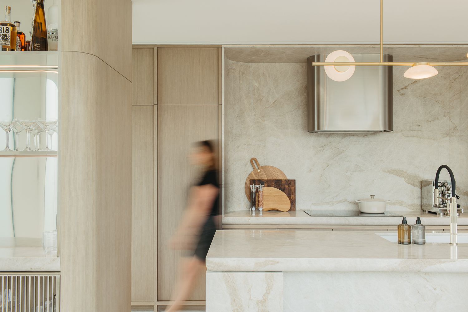 Elegant modern kitchen with marble countertops, beige cabinetry, pendant lights, and a blurred figure walking past.