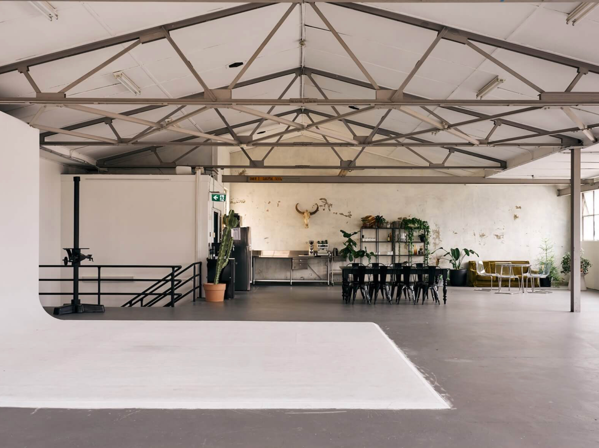 Spacious industrial photography studio with high truss ceiling, white backdrop, dining area, and lush plants.