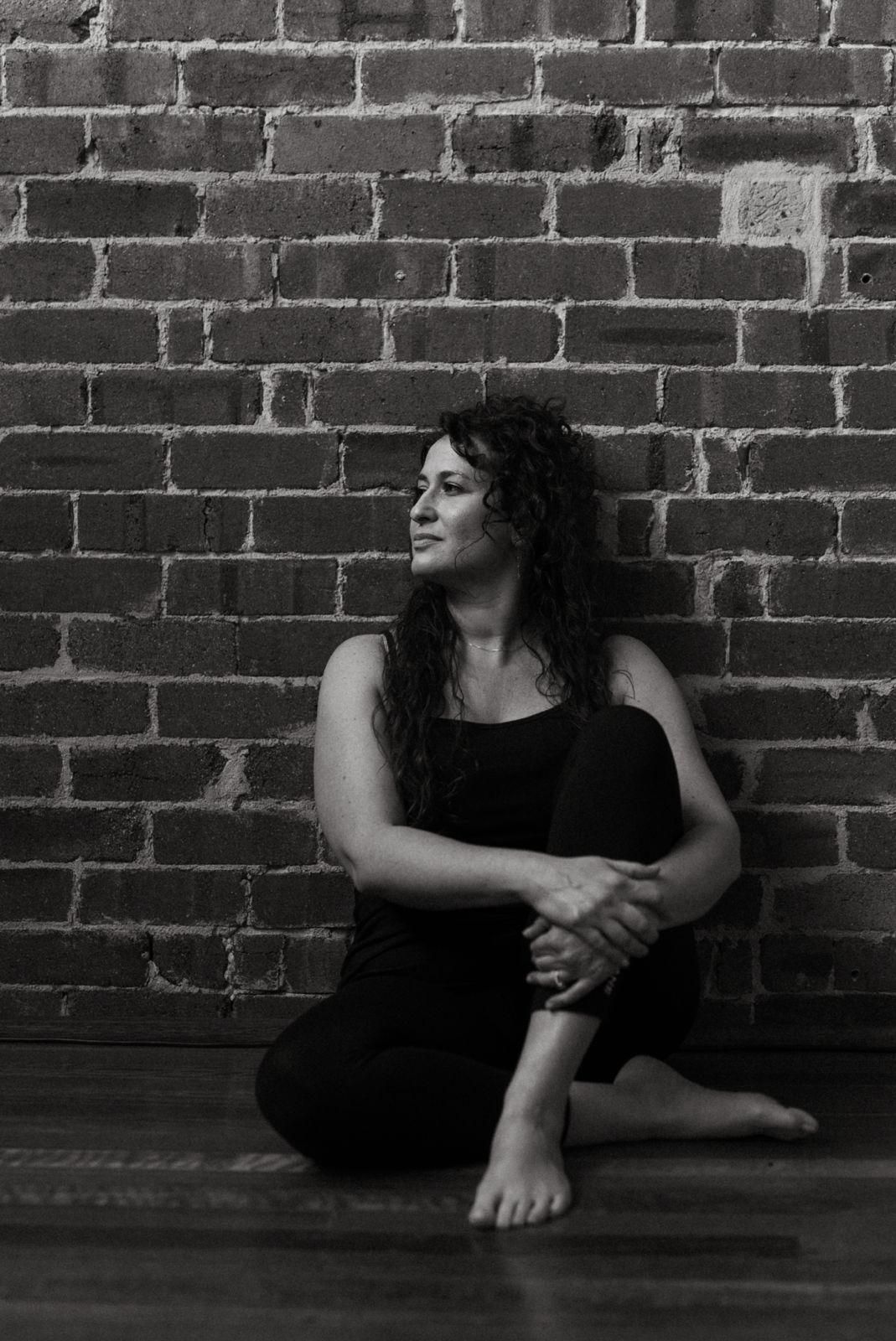 Black and white photo of a woman with curly hair sitting cross-legged on a wooden floor against a brick wall.