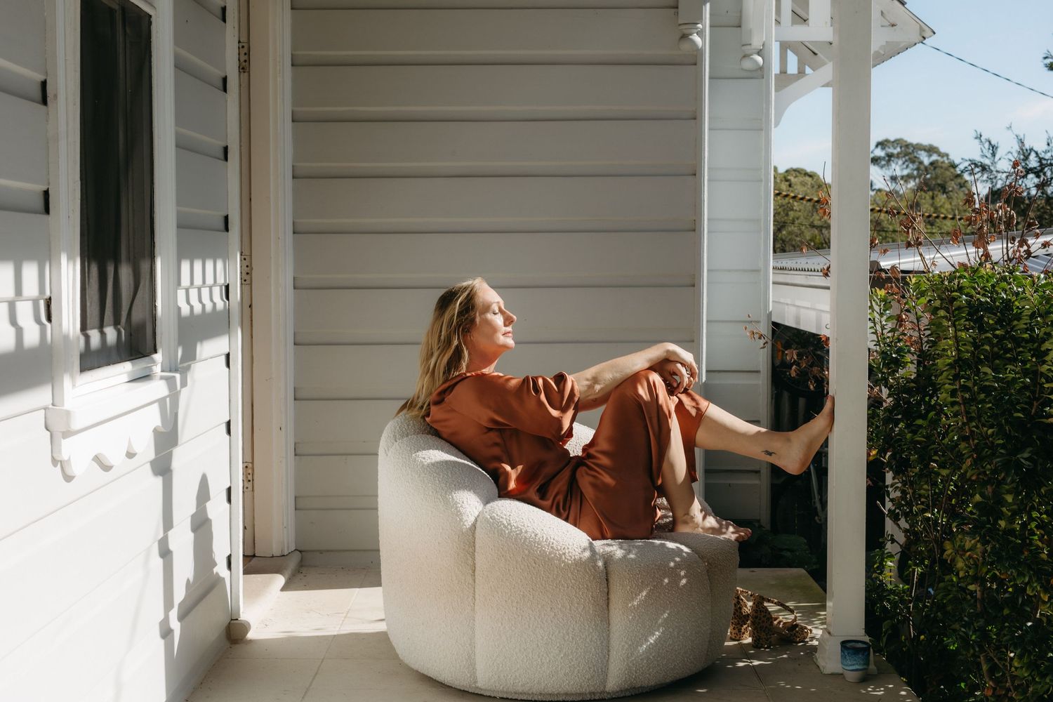 Woman relaxing in a white bean bag chair on a sunny porch wearing a rust-colored outfit.