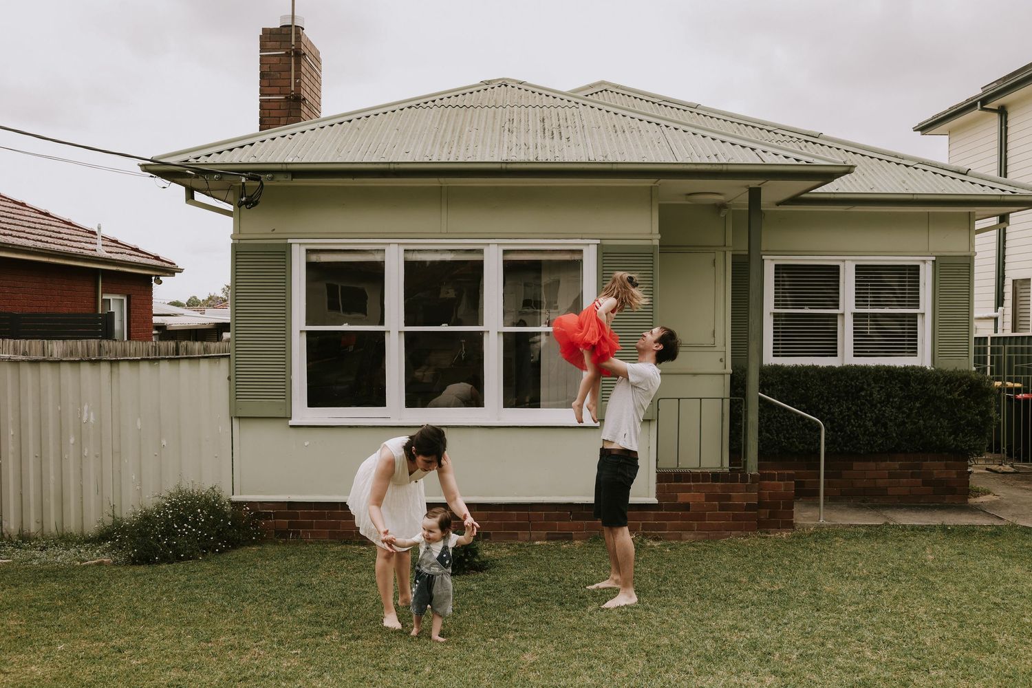 Family with dog and child playing with red balloons in front of a green suburban house on a cloudy day.