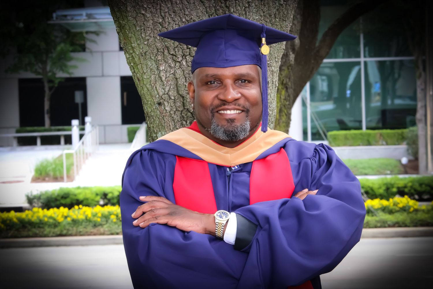 Man in blue and red graduation cap and gown smiling with arms crossed outdoors.