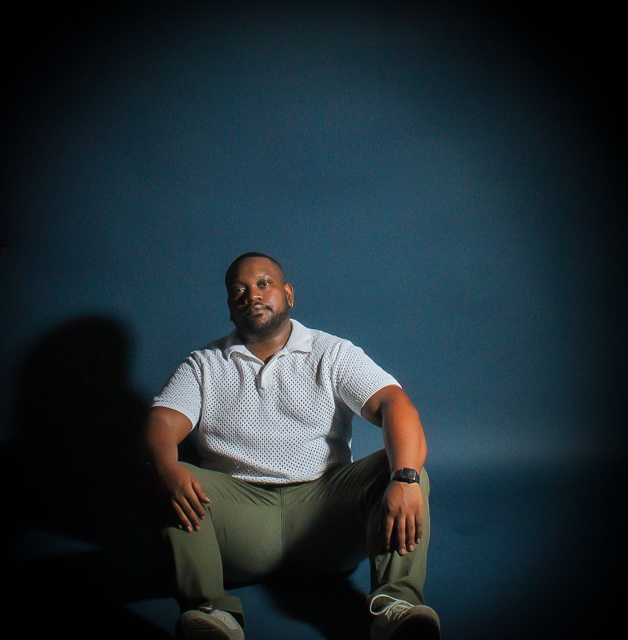 Man in white polo shirt and khaki pants sitting confidently against a dark blue background in dramatic lighting.