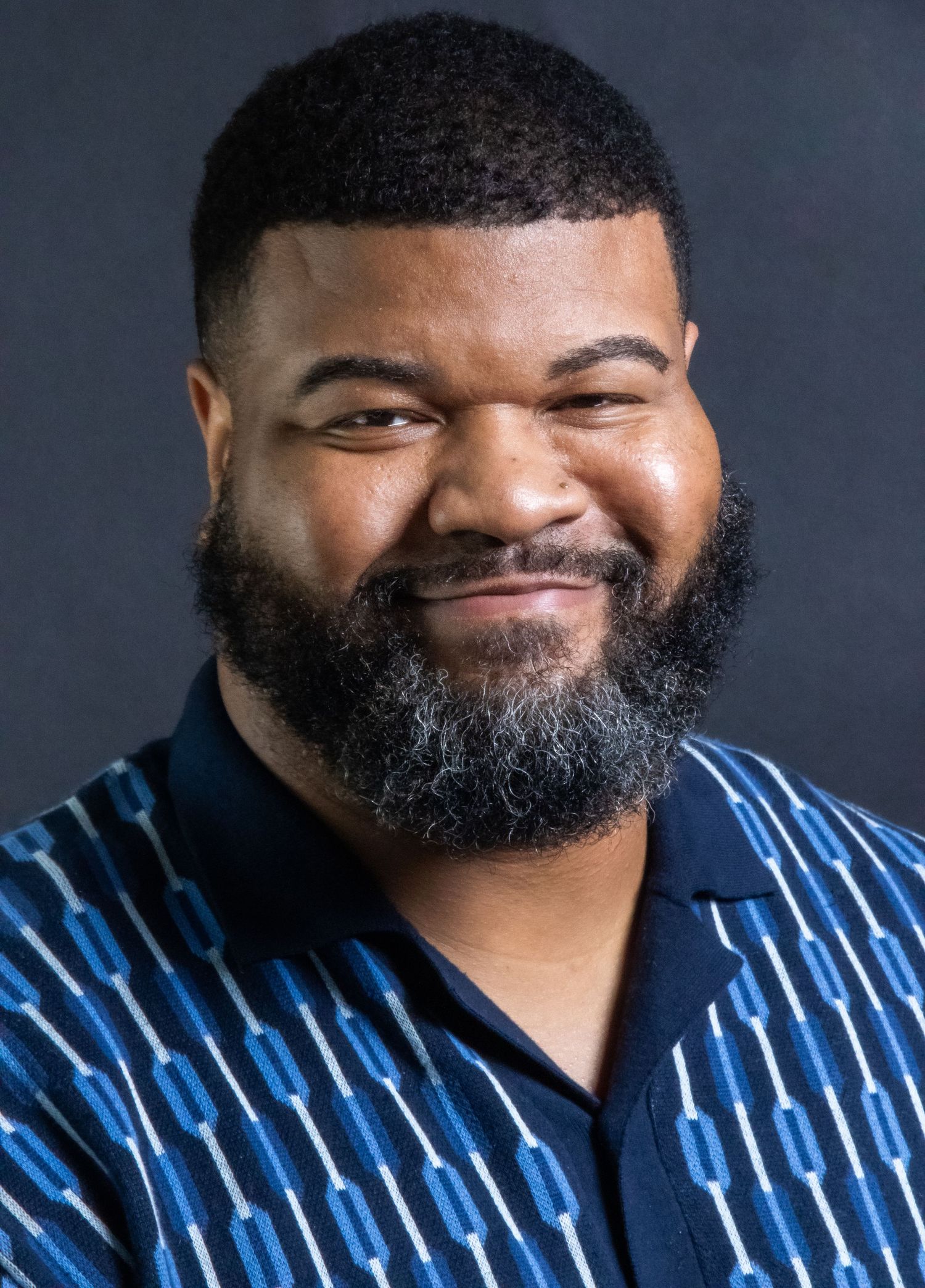 Smiling Black man with beard wearing a blue patterned polo shirt poses for a professional headshot photo.