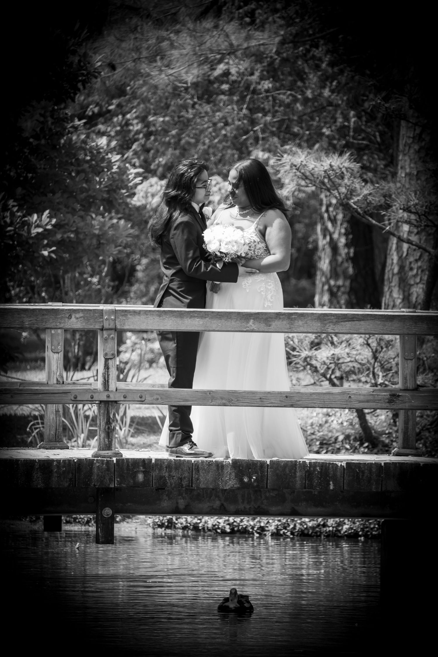 Black and white photo of a bride and groom sharing a romantic moment on a wooden bridge over a pond.