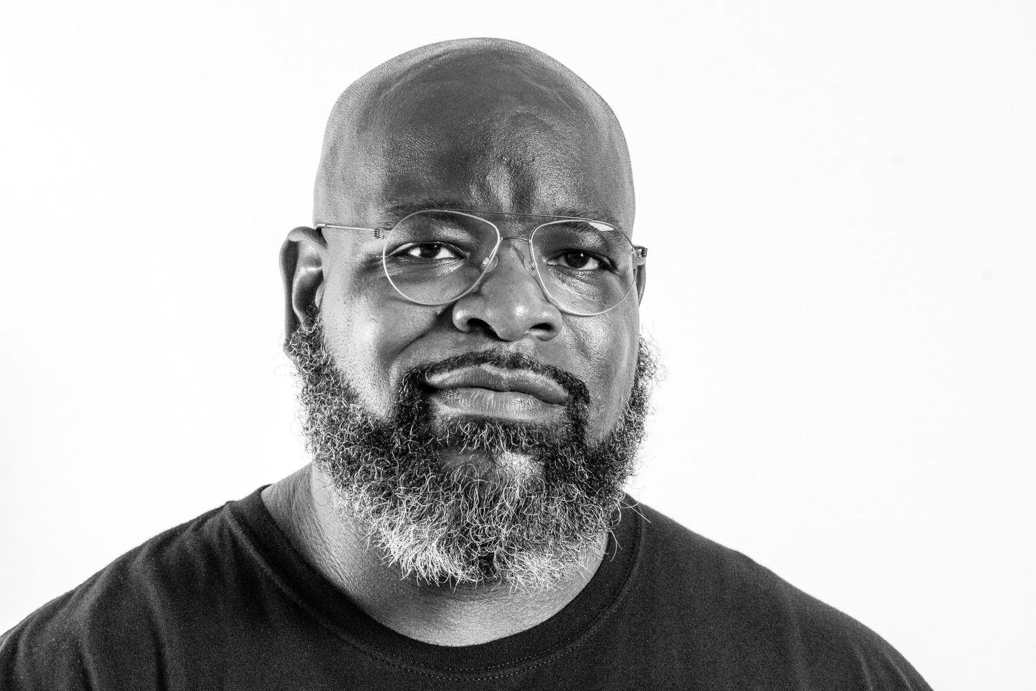 Black and white portrait of a bald man with a beard and glasses wearing a black t-shirt.