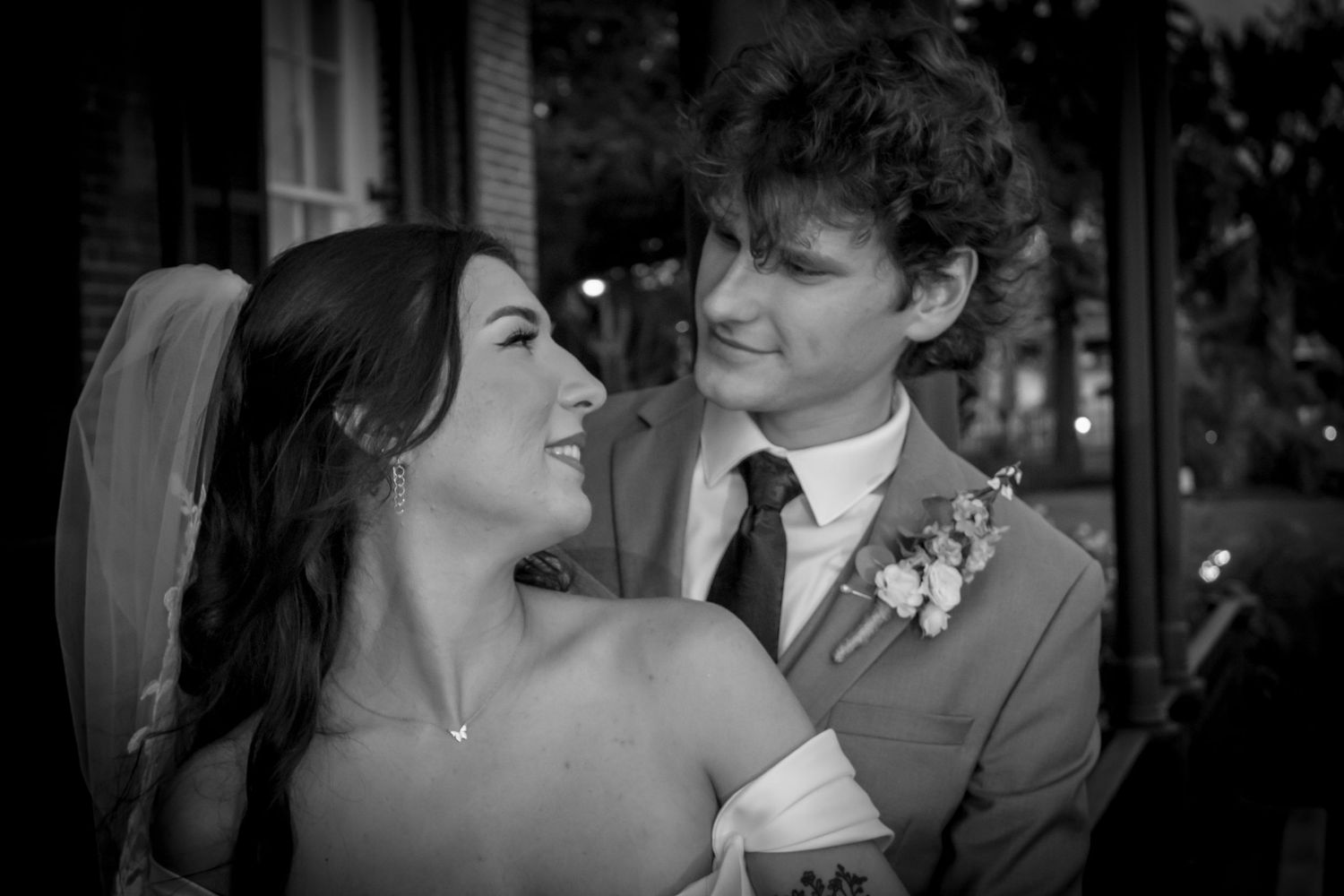 Black and white wedding photo of smiling bride and groom in suit with boutonniere gazing at each other romantically.
