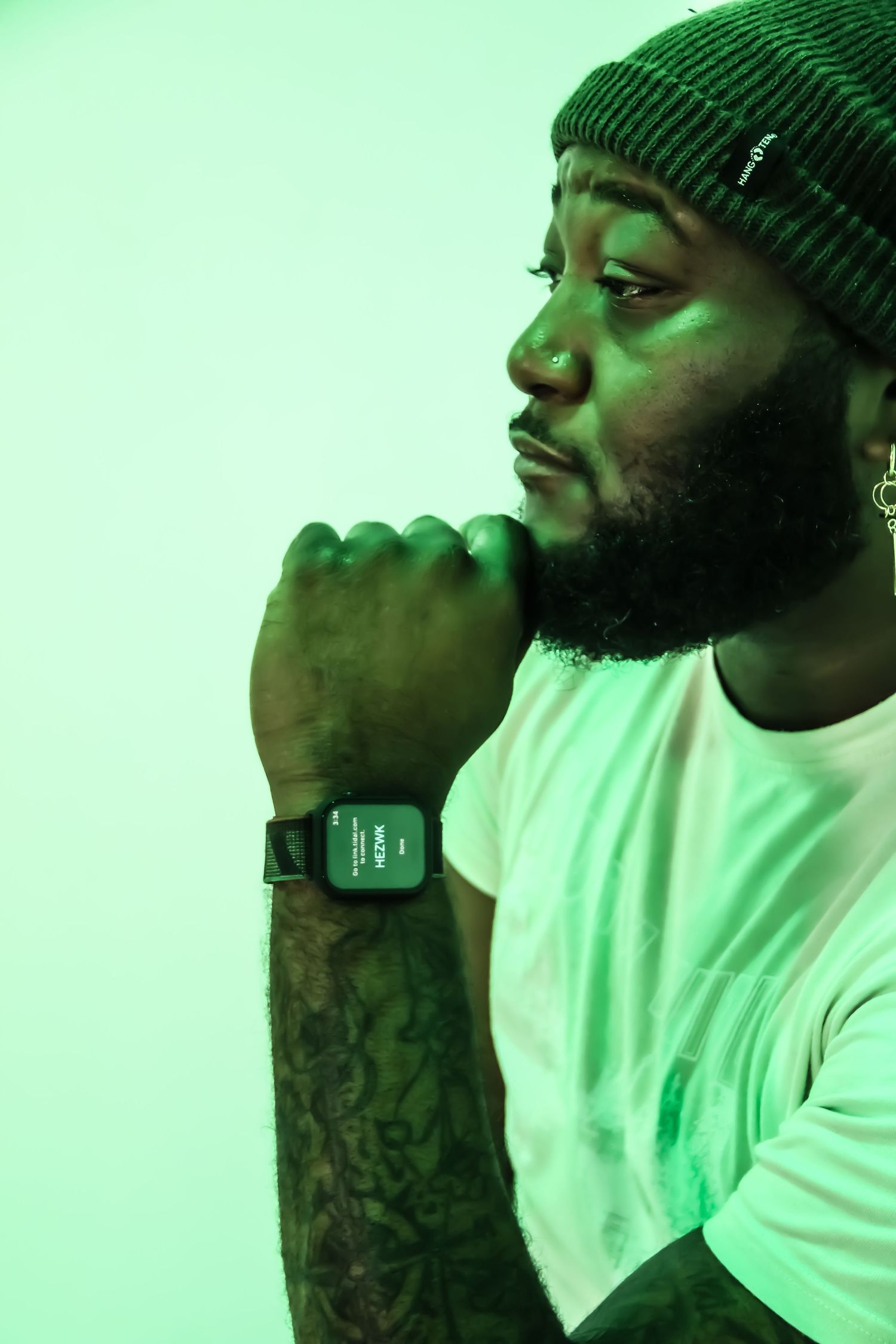 Bearded Black man in green beanie and white tee with tattoos poses thoughtfully under green neon light wearing a smartwatch.