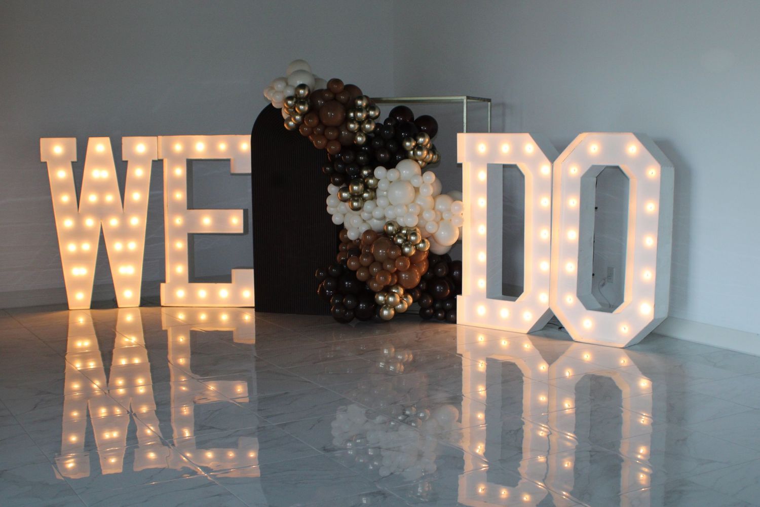 Illuminated marquee WE DO letters with balloon garland backdrop for wedding decor.