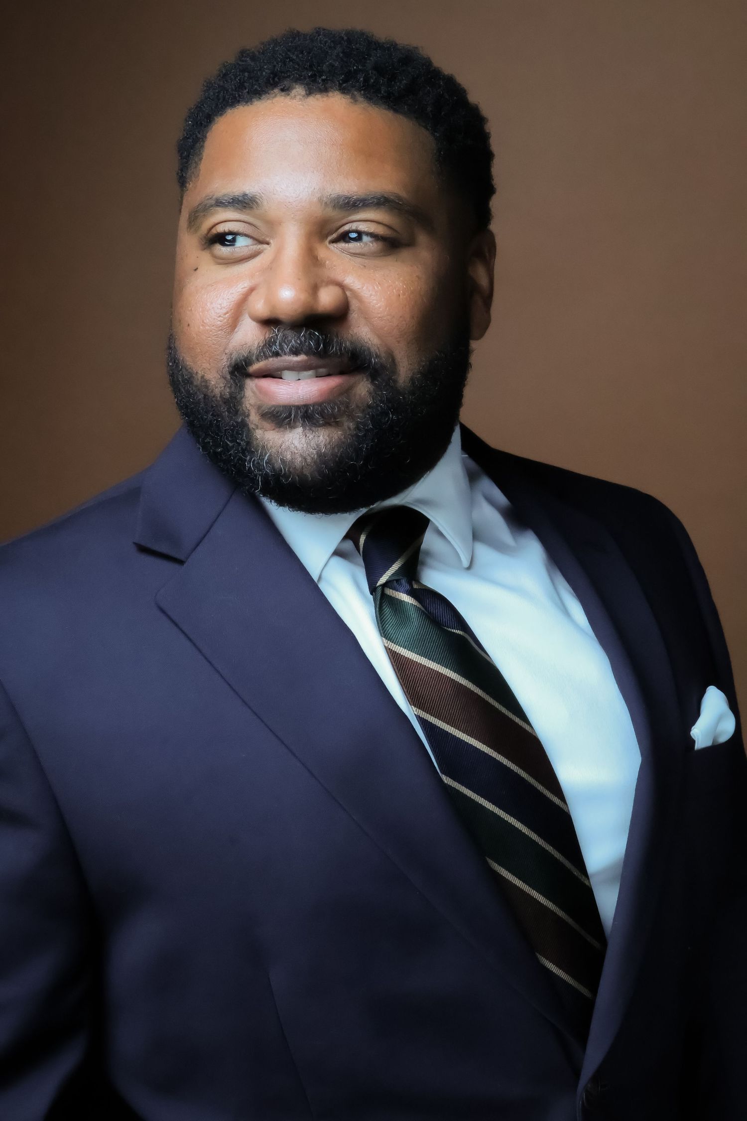 Professional headshot of a bearded man in a navy suit and striped tie against a brown background.