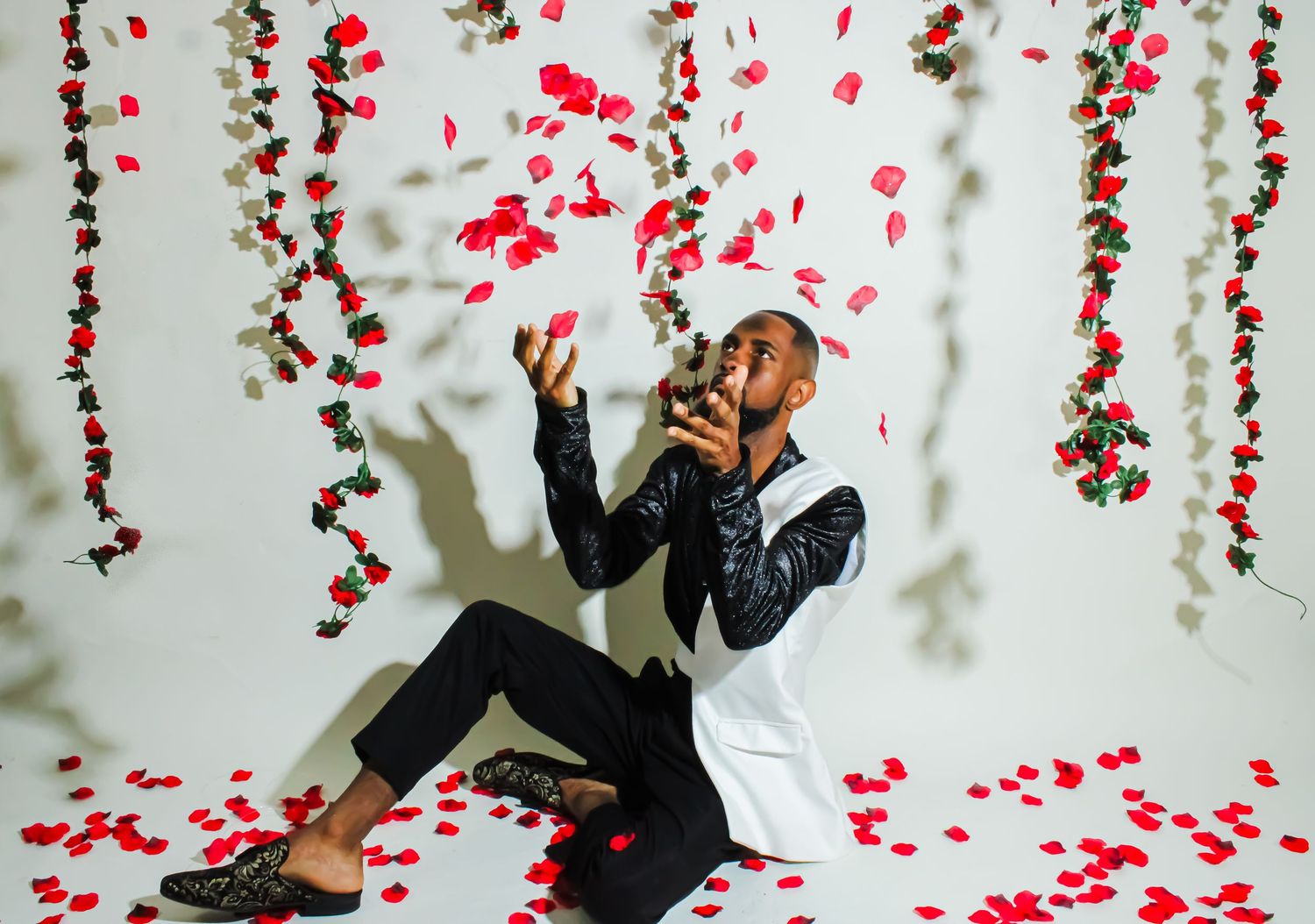 Man in white suit tossing red rose petals in air against white backdrop with hanging floral garlands.