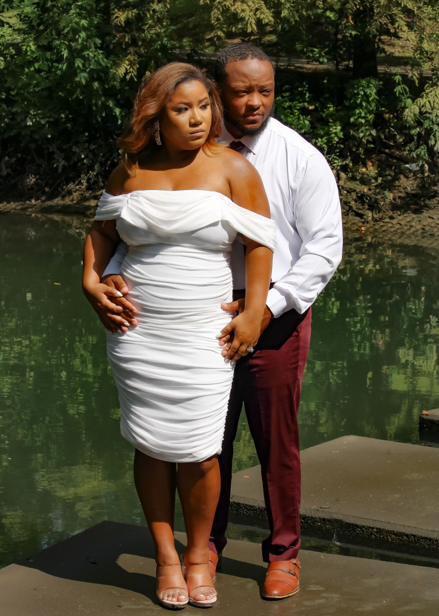 Couple posing outdoors near a pond, woman in white off-shoulder dress, man in white shirt and burgundy pants.