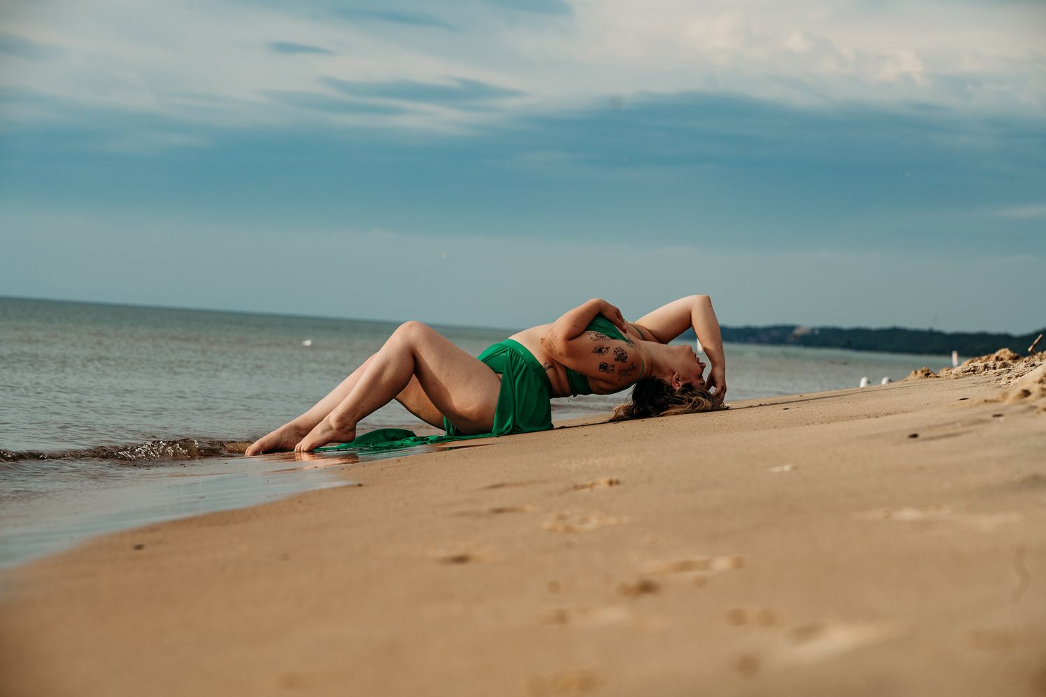 Woman in green bikini lying on sandy beach shore with calm ocean waves and cloudy blue sky in background.