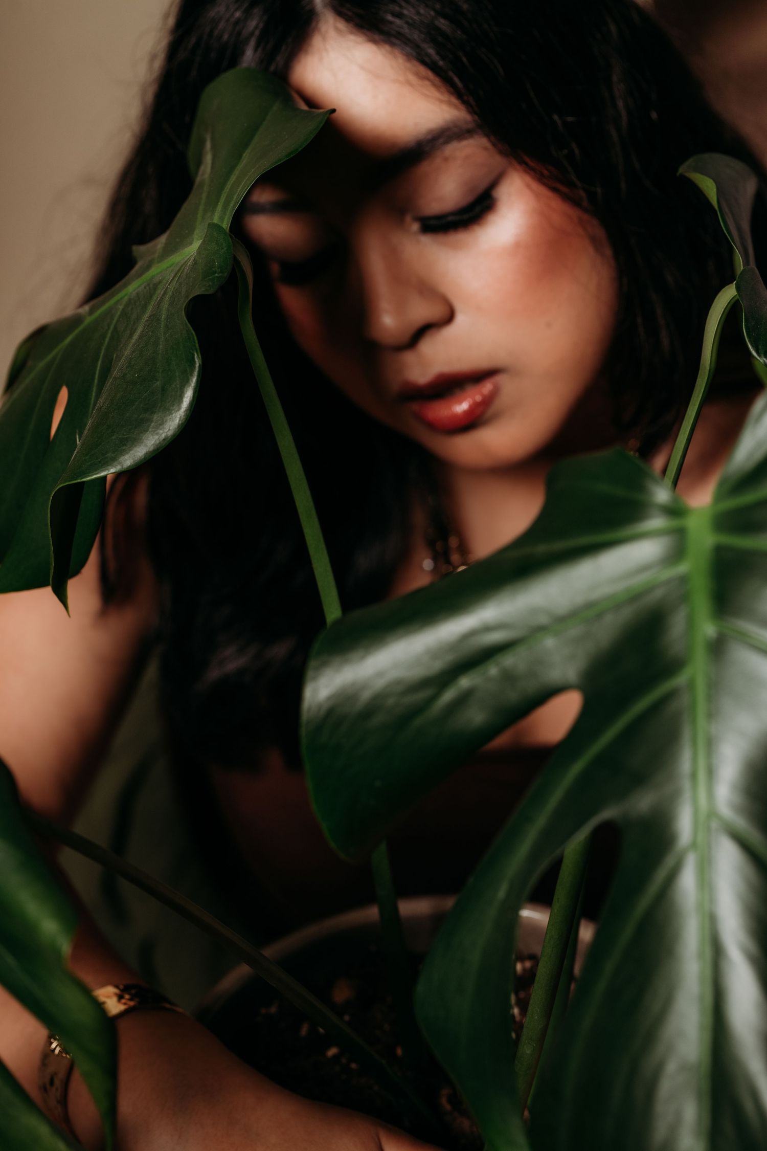 A young woman with long dark hair poses gracefully among large green monstera leaves in a moody, artistic portrait.