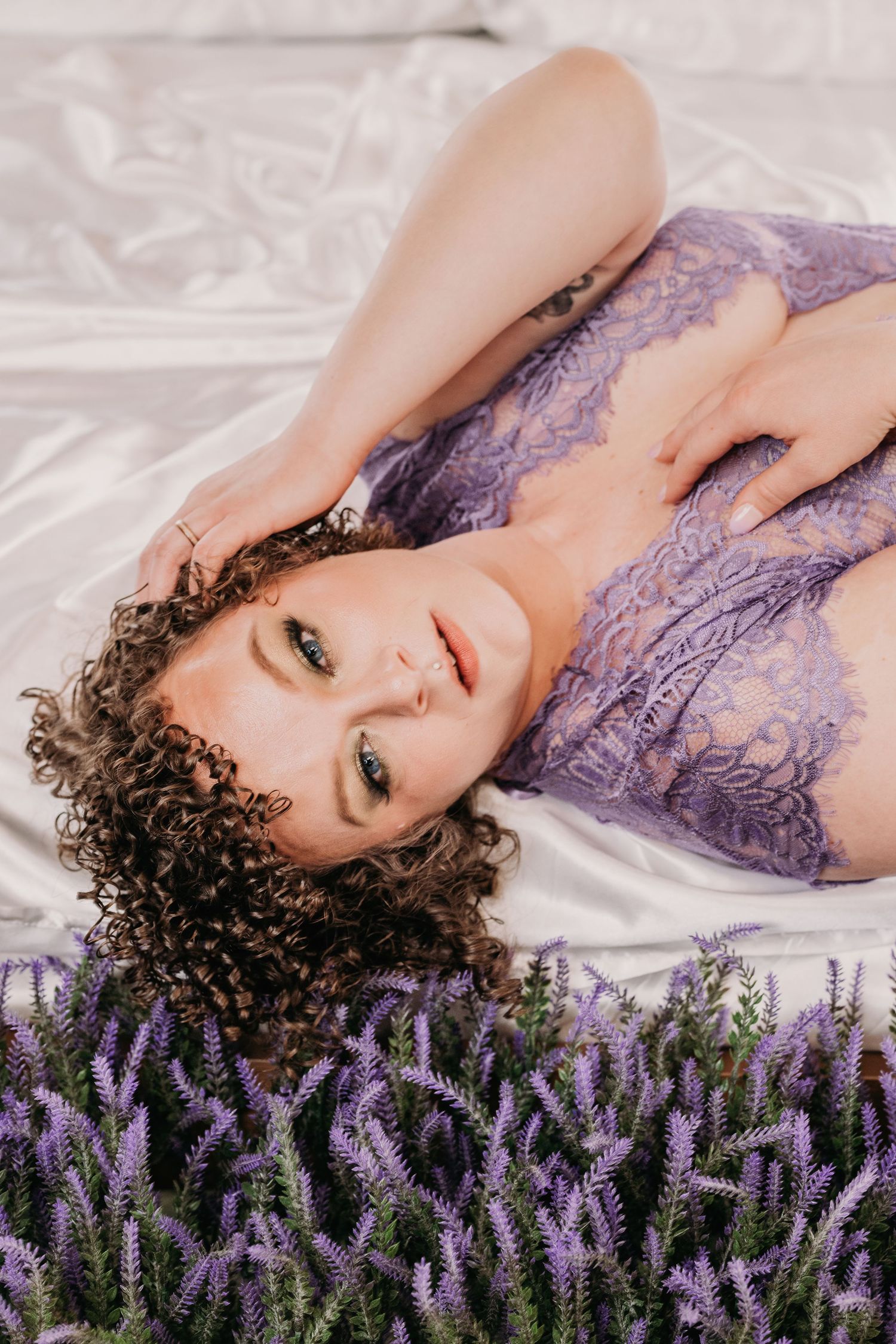 Boudoir photoshoot with lavender flowers on white sheets featuring delicate purple lace lingerie. Boudoir Photography Studio, Grand Rapids M