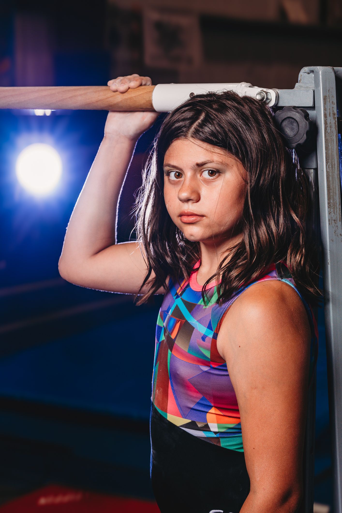 Young female gymnast in colorful leotard gripping a horizontal bar in a dimly lit gymnasium.