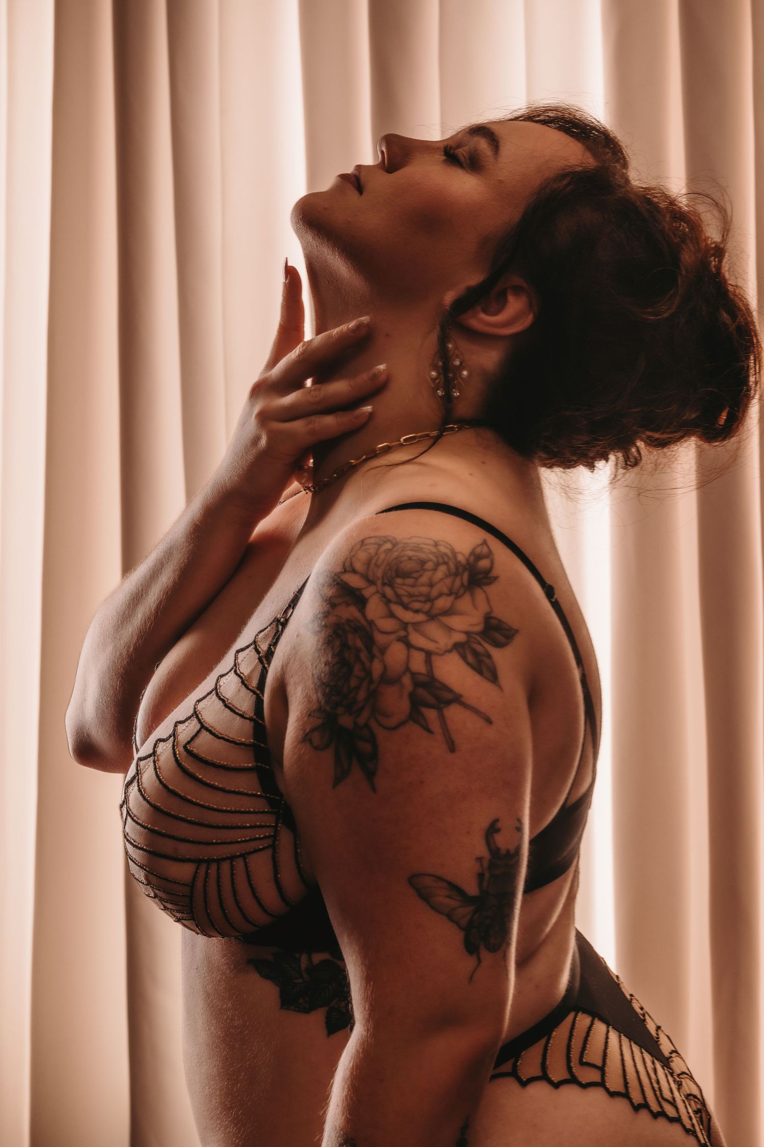 Artistic portrait showing butterfly and floral tattoos in warm sepia tones against beige curtained background. Bridal Boudoir Photography