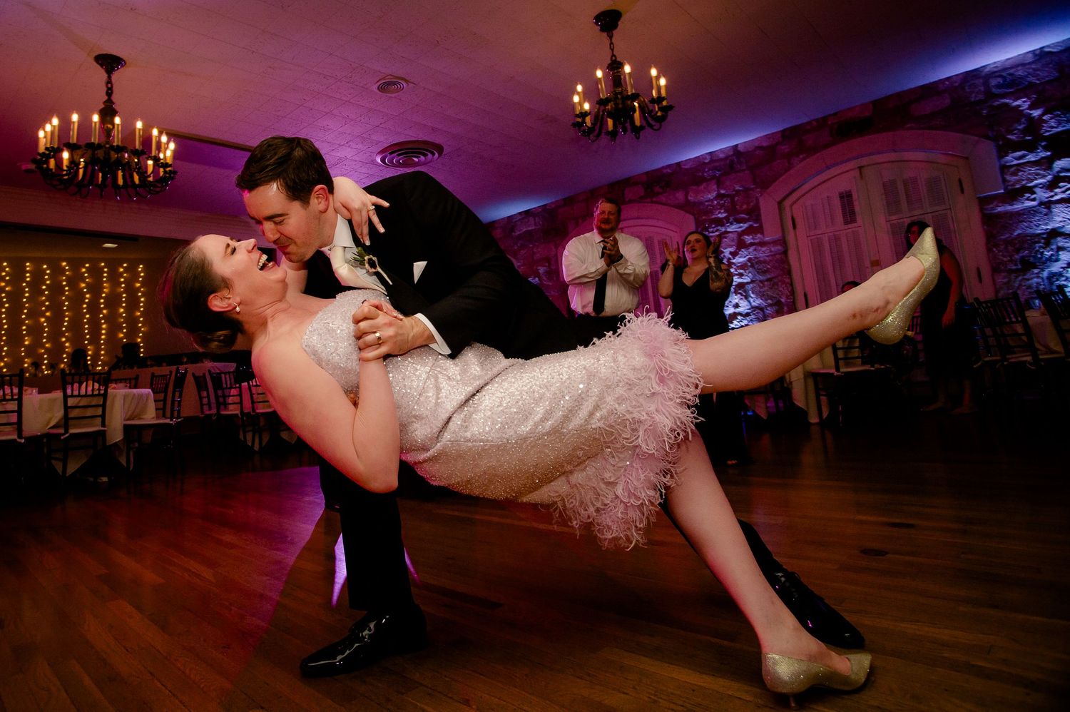 Bride and groom share romantic dip dance at wedding reception in elegant ballroom with purple lighting.