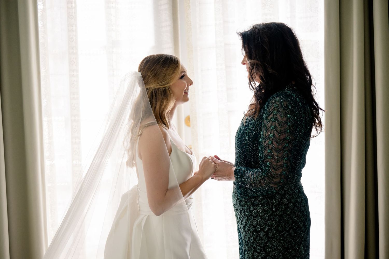 Bride in white gown and veil holds hands with woman in green lace dress, sharing a tender moment by a bright window.